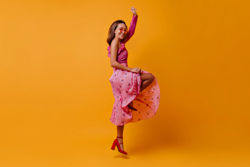 happy, affable female model in pleated long skirt bounces, showing graceful slender legs in unusual shoes. Brunette with glasses has fun in studio