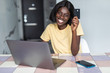 © F8  \ Suport Ukraine - Pretty young african woman using laptop and holding credit card in kitchen