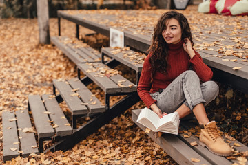 Beautiful woman in cozy outfit reading book sitting on wooden bench in autumn park.