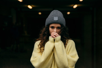 Young curly  girl in yellow sweater and gray cap stay alone at the city lights and freeze