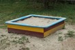 colored square wooden sandbox with white sand stands on the ground and green grass in the playground outside