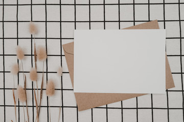 Blank white paper on brown paper envelope with Rabbit tail dry flower and Carton box on Black cloth with Black White grid pattern. Mock-up of horizontal blank greeting card. Top view of Craft envelope