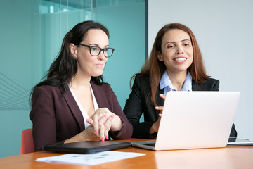 Businesswomen having video talk to partners, sitting at open laptop, looking at display and smiling. Medium shot. Business communication concept