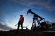 Horizontal snapshot of a silhouette of an oilman in orange vest and helmet, standing with his back to the oil pump jack in oilfield holding a pipe wrench, looking away, evening sky on background
