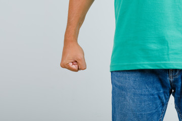 Young man standing with clenched fist in green t-shirt, jeans and looking angry , front view.