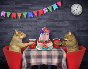 Wall Mural - A cat with a dog together are eating a holiday two tiered cake and drinking coffee at a table in a restaurant.