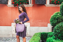 Young Beautiful Brunette Woman Wearing Purple Polka Dot Dress, Holding Knitted Bag With Flowers, Posing In Street Of City. Copy, Empty Space For Text