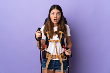 Young woman with backpack and trekking poles isolated on purple background with surprise and shocked facial expression