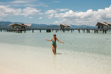 Wall Mural - Young slim tourist woman enjoy suntan, standing in the tourquise water in front of wooden owrwater bungalows, wearing swimsuit. Vacation concept