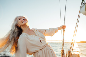 Wall Mural - Beautiful mature woman with long hair enjoying sunset. Smiling woman holding a rope and looking up while standing on a sailboat.