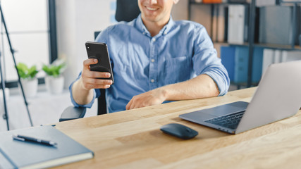 Poster - Creative Entrepreneur Sitting at His Desk Works on a Laptop in the Stylish Office, Uses Smartphone for Social Media Apps, Emailing Business Associates, Reading News, Browsing through Internet
