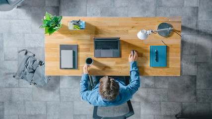 Poster - Top View Shot: Businessman Sitting at the Wooden Desk Works on a Laptop in his Home Office. He Drinks Coffee, Types, Writes Emails, Surfs the Internet, Designs Software, Online Shopping