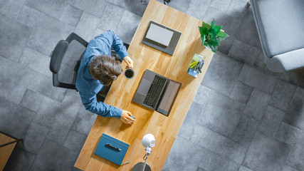 Poster - Top View Shot: Businessman Sitting at the Wooden Desk Works on a Laptop in his Home Office. He Drinks Coffee, Types, Writes, Surfs the Internet, Creates and Designs Software, Online Shopping