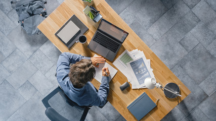 Poster - Businessman Sitting at His Desk in the Office Uses Laptop Computer, Working with Company Strategy Documents, Correcting Charts, Drawing Graphs and Statistics. Top View Shot