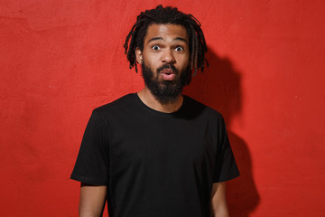 Shocked amazed surprised young african american man guy with dreadlocks 20s wearing black casual t-shirt posing saying wow looking camera isolated on bright red color wall background studio portrait.