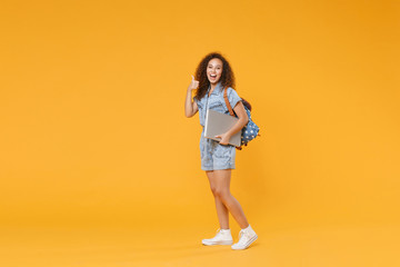 Wall Mural - Full length portrait funny african american girl student in denim clothes, backpack isolated on yellow background. Education in high school university college. Hold laptop computer, showing thumb up.