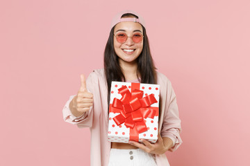 Wall Mural - Smiling young asian woman in casual clothes cap glasses isolated on pink background. Valentine's Day Women's Day birthday, holiday concept. Hold red present box with gift ribbon bow, showing thumb up.