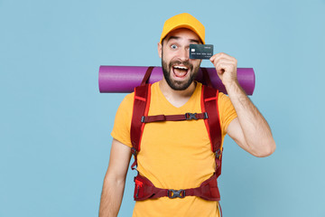 Wall Mural - Cheerful traveler young man in yellow casual t-shirt backpack isolated on blue background. Tourist traveling on weekend getaway. Tourism discovering hiking concept. Covering eye with credit bank card.