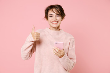 Smiling young brunette woman girl wearing knitted casual sweater posing isolated on pastel pink wall background studio portrait. People lifestyle concept. Using mobile cell phone, showing thumb up.