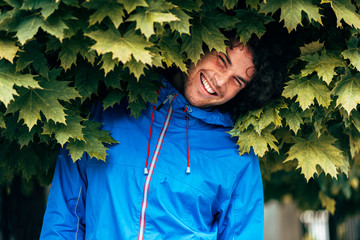 Wall Mural - Outdoor portrait of a happy curly young man smiling broadly, looking to the camera wearing blue raincoat during the rain outside. Handsome male enjoying the rainy day hiding in the green leaves.