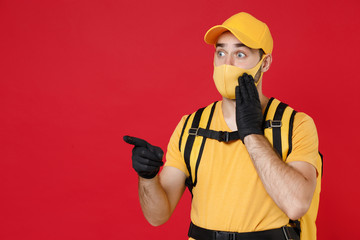 Wall Mural - Delivery employee man in yellow cap face mask gloves t-shirt uniform thermal bag backpack with food work courier service during quarantine coronavirus covid-19 virus isolated on red background studio.