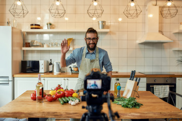 Young man, Italian cook in apron waving at camera, greeting audience, filming himself for culinary blog while preparing healthy meal with vegetables in the kitchen