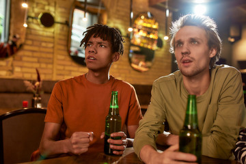 Friends in the bar looking disgruntled while watching sports match on TV together, drinking beer and cheering for team. People, leisure, friendship and entertainment concept