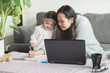 © Carlos Lopez - Asian woman working from home helps her daughter draw on a notepad while working on her laptop computer, sitting on a sofa in the living room of a house in Edinburgh, Scotland, UK, as both smile