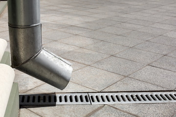 street engineering structure a downspout pipe with drainage gutter on a background of a gray stone granite sidewalk from square tiles with copy space, side view closeup nobody.