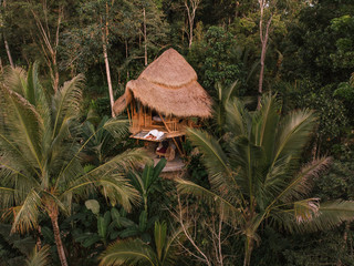 Aerial view from drone happy travel couple on hammock balcony of bamboo tree house with jungle nature view. Vacation in beaitiful hidden place, honeymoon on Bali island