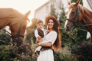 a mother in a hat with her son in her arms stands next to two beautiful horses in nature. a family with a child is photographed with horses