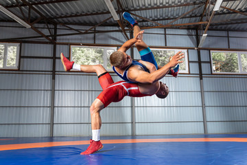 Two  strong men in blue and red wrestling tights are wrestlng and making a suplex wrestling on a yellow wrestling carpet in the gym. Wrestlers doing grapple.