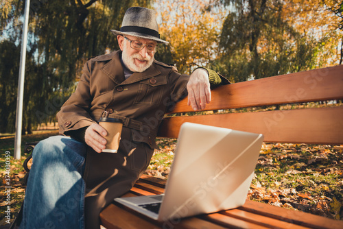 Photo of white grey hair old man enjoy october fall rest nature park relax sit bench watch modern movie laptop hold drink takeaway coffee mug wear cap hat headwear coat jacket