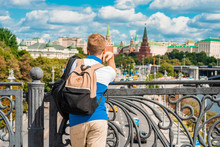 A Young Blond Man Stands On A Bridge And Admires The Kremlin And The River In Moscow, Photo From The Back