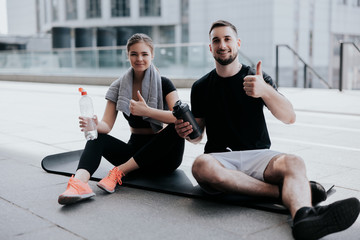 Wall Mural - happy young couple brunette beautiful woman and handsome man in sportswear show like sign, stretching on yoga mat, drinking water , healthy fit lifestyle, street workout