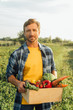 © LIGHTFIELD STUDIOS - farmer in plaid shirt looking at camera while holding box with fresh, ripe vegetables