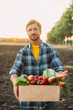© LIGHTFIELD STUDIOS - selective focus of farmer in plaid shirt holding wooden box with ripe vegetables while looking at camera