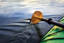 Boat Oar And Part Of Green Kayak In The Water And Splashing Water At Evening River At Sunset