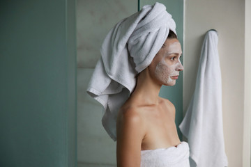 Young woman in bathroom wrapped in towels with facial mask. Home spa and skin care concept.