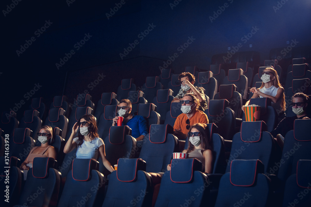 Cinema, movie theatre during quarantine. Coronavirus pandemic safety rules, social distance during movie watching. Men and women wearing protective face mask sitting in a rows of auditorium, eating - obrazy, fototapety, plakaty 