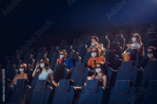 Cinema, movie theatre during quarantine. Coronavirus pandemic safety rules, social distance during movie watching. Men and women wearing protective face mask sitting in a rows of auditorium, eating