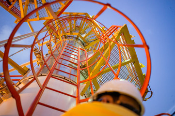 Bottom up pov of oil worker on an industrial steel ladder with safety cage. Heavy industry gas and petroleum plant. Pov with selective focus and vivid yellow, orange and reflective steel colors as saf