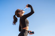© DimaBerlin - Thirsty young fit woman drinking water from plastic bottle, resting after jogging, holding mobile smartphone in her hand, listens to music. Blue sky with copy space. Hot day.