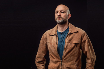 Portrait of a bald man in his 40s dressed in brown suede jacket. Model with grey and beard. Dark background.