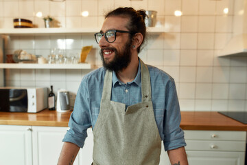 Portrait of young man, Italian cook in apron smiling aside while getting ready to prepare healthy meal with vegetables in the kitchen. Cooking at home concept