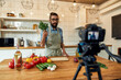 © Svitlana - Young man, Italian cook in apron looking at camera, holding basil, filming himself for culinary blog while preparing healthy meal with vegetables in the kitchen