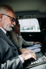 Wall Mural - Good looking senior business man and his young woman colleague or coworker sitting on backseat in luxury car. They talking, smiling and using laptop and smart phones. 