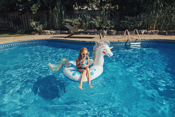 Wall Mural - Cute adorable girl in sunglasses with drink lying on inflatable ring unicorn. Kid child enjoying having fun in swimming pool. Summer outdoors water activity for kids.