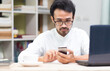 © Surasak Chuaymoo - Young man working on mobile phone application at  office desk in busy  with laptop and coffee and book shelf background, Working connection concept