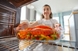 culinary, food and people concept - woman cooking salmon fish with vegetables in baking dish in oven at home kitchen